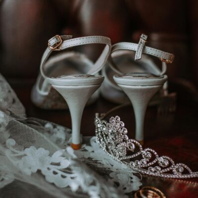 Elegant bridal high heeled shoes and veil with golden rings and tiara placed on leather sofa before wedding celebration