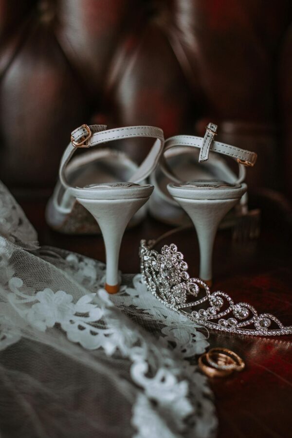 Elegant bridal high heeled shoes and veil with golden rings and tiara placed on leather sofa before wedding celebration