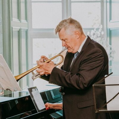 Side view of concentrated senior musician with grey hair in dark elegant suit looking at notes on sheet and playing trumpet next to pianist
